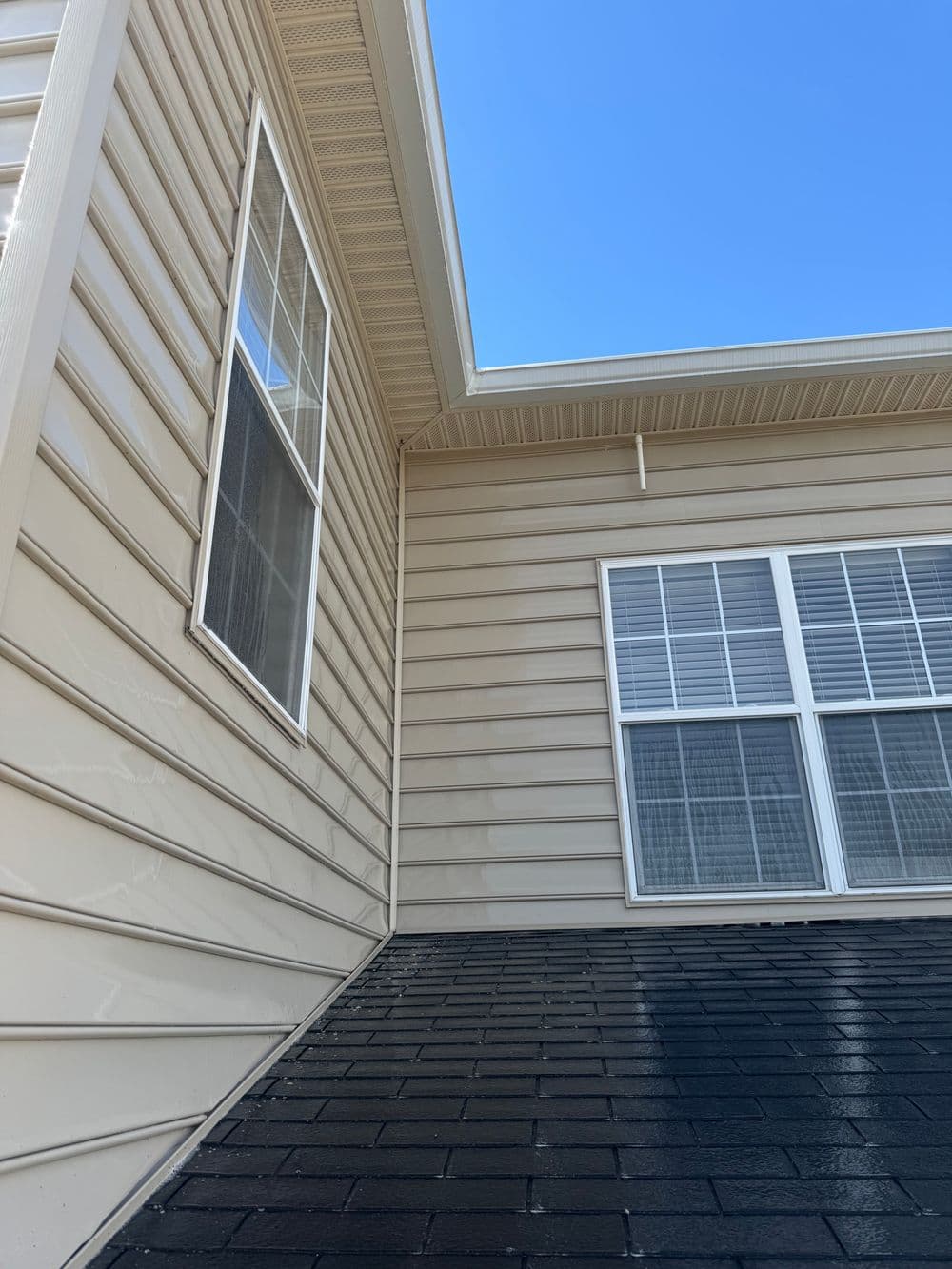 Close-up of a house corner showing beige siding and dark shingles under a clear blue sky.
